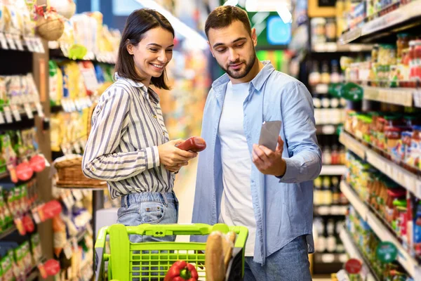 Young couple with cart and smartphone shopping in supermarket