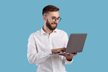 Resmi giyinen başarılı genç adam mavi stüdyo üzerinden dizüstü bilgisayar kullanıyor.