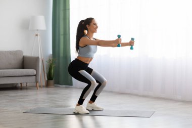 Atletik genç bayan evde dambıllarla çömelme yapıyor.