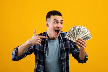 Heyecanlı genç adam turuncu stüdyo arka planında elinde para yelpazesini işaret ediyor.