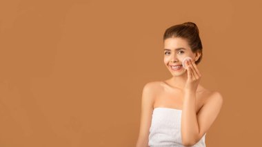 Güzel genç bayan yüzünü pamukla siliyor, kahverengi arka planda su veya tonikle cildini temizliyor.