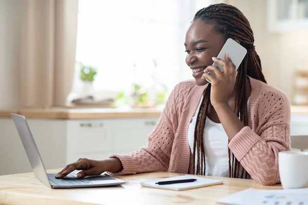 Telecommuting Concept. Black Woman Using Cellphone And Working On Laptop In Kitchen