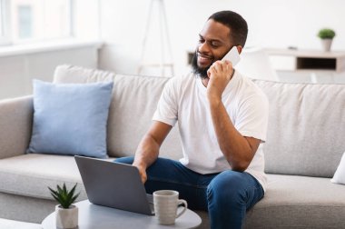 Kapalı alanda dizüstü bilgisayarda çalışırken cep telefonuyla konuşan adam.