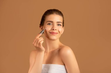 İpeksi ciltli güzel bir kadın yüz kremi sürüyor ya da kahverengi stüdyo arka planında kozmetik fosforlu kalem kullanıyor.