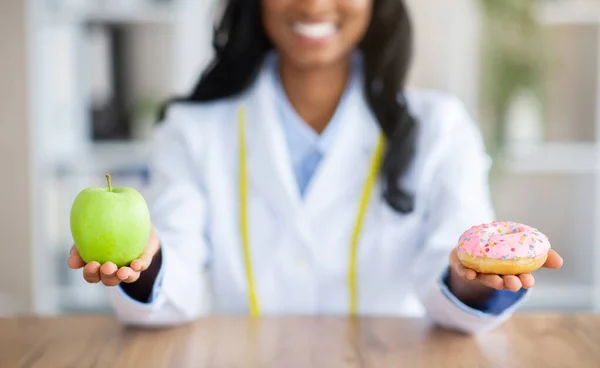 Vue rapprochée des mains des diététistes tenant une pomme fraîche et un beignet à table dans un cabinet médical. Concept de régime — Photo
