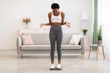 Memnuniyetsiz siyah kadın terazinin üzerinde duruyor evde tartıyor kendini