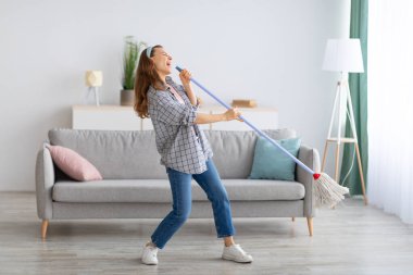 Mutlu genç ev hanımı mikrofon olarak paspas kullanıyor, ev işleri sırasında şarkı söylüyor, kapalı alanda.