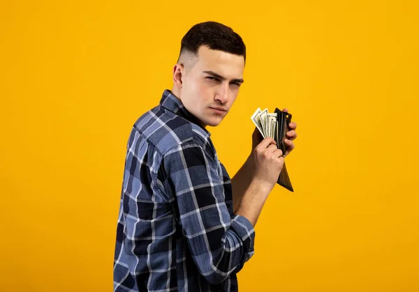 Fiatal férfi pénztárcával tele pénzt, hogy kapzsi a vagyonát narancssárga stúdió háttér — Stock Fotó