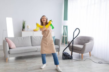 Neşeli genç hizmetçi elinde deterjan şişeleriyle temiz bir evde silah gibi, boş bir yerde.