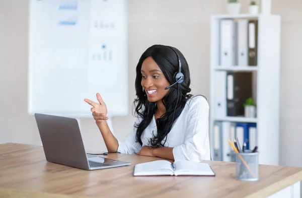 Koncepcja telekomunikacji. Uśmiechnięta Afroamerykanka ze słuchawkami pracująca online na laptopie w biurze — Zdjęcie stockowe