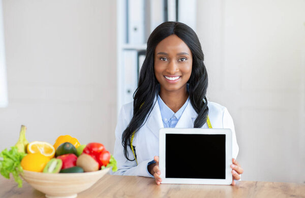 Healthy nutrition. Happy black dietitian showing tablet computer with blank screen at table in her office, mockup
