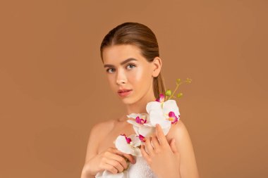 Orkide çiçeği tutan güzel genç bir bayan ve kahverengi stüdyo arka planındaki kameraya bakıyor. Güzellik ve sağlık.
