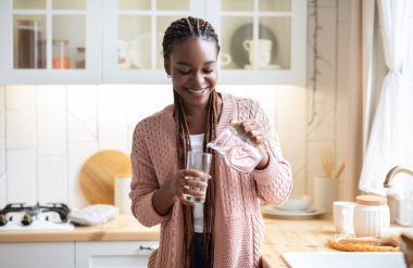 Susamış siyah kadın mutfakta su içiyor, bardağa sağlıklı sıvı döküyor.