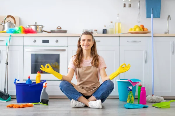 Happy millennial housewife sitting in lotus yoga pose on kitchen floor surrounded by cleaning supplies