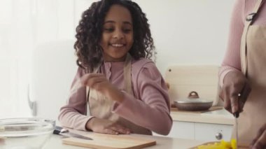 Birlikte yemek pişirmek. Anne mutfakta sebze kesiyor, küçük tatlı bir kız bir parça alıyor ve gülümsüyor.