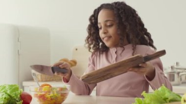 Küçük şef. Afro-Amerikalı kız öğle yemeğini kendisi hazırlıyor, sebze doğruyor ve mutfakta salata karıştırıyor.
