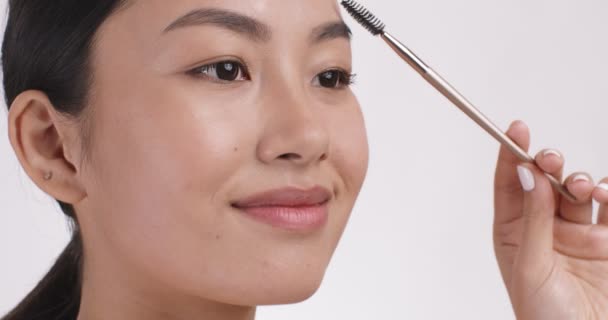 Jong aziatisch dame styling haar wenkbrauwen met wenkbrauw mascara, wit studio achtergrond, close up, slow motion — Stockvideo