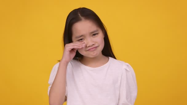 Проблемы детей. Портрет расстроенной азиатской девушки, плачущей и вытирающей слезы, оранжевый фон, замедленная съемка — стоковое видео