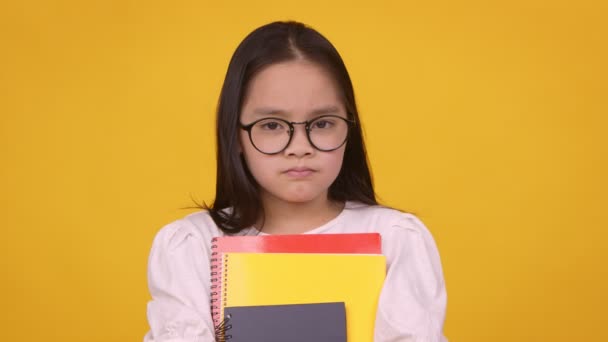 Pequeña colegiala asiática en gafas sosteniendo libros de texto y sacudiendo la cabeza no, fondo de estudio naranja — Vídeos de Stock