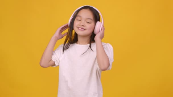 Aranyos kis ázsiai lány élvezi a klasszikus zene és tánc, visel vezeték nélküli headset, narancs háttér, lassított felvétel — Stock videók
