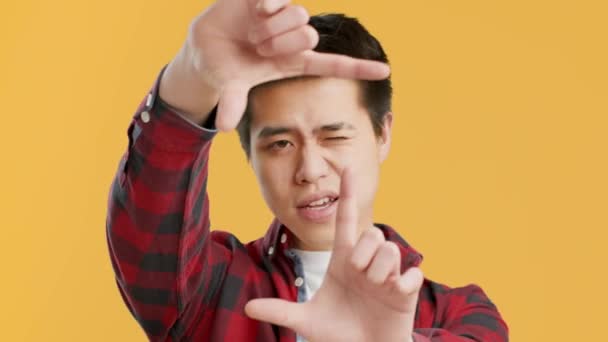 Азиатский парень ловит момент, обрамляя лицо пальцами, желтым фоном — стоковое видео