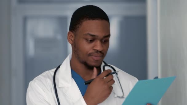 Médico masculino afro-americano atencioso olhando para os resultados do teste e pensando, segurando prancheta no hospital, close-up — Vídeo de Stock