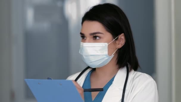 Papeleo en el hospital. Médico femenino positivo en la escritura protectora de la máscara médica en el portapapeles, teniendo gran idea — Vídeo de stock