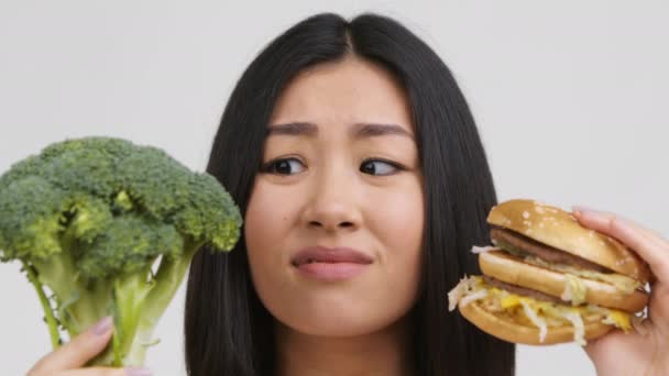 Mulher asiática escolhendo hambúrguer não saudável em vez de brócolis, fundo branco — Vídeo de Stock