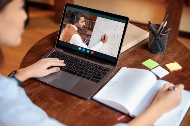 Masada oturan kadın dizüstü bilgisayar kullanıyor, öğretmenle görüntülü konuşma yapıyor.