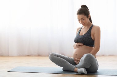Evde yoga minderinde oturan spor kıyafetli güzel hamile kadın.