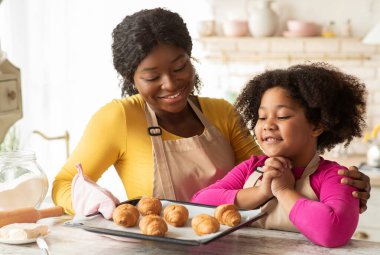 Küçük siyah kız mutfakta yeni pişmiş kruvasanlarla tepsiye bakıyor.