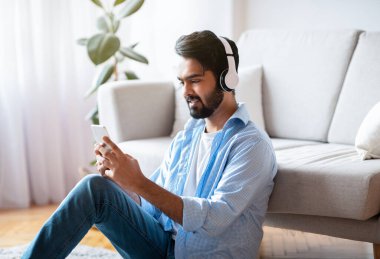 Yakışıklı Arap Adam, evde akıllı telefon ve kablosuz kulaklıkla müzik dinliyor.