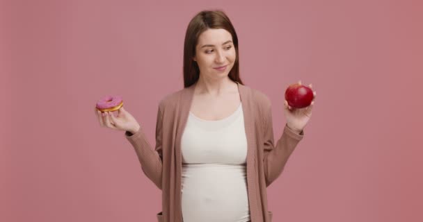 Estúdio tiro de jovem grávida comparando donut doce e maçã madura e ombros encolhendo na incerteza — Vídeo de Stock