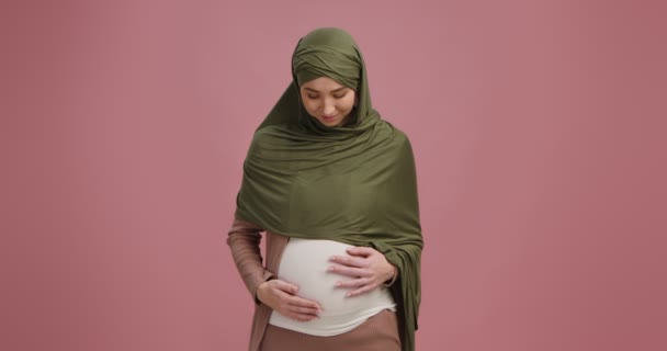 Νεαρή μουσουλμάνα έγκυος με χιτζάμπ χαϊδεύει τη μεγάλη κοιλιά της, απολαμβάνει την εγκυμοσύνη της, ροζ φόντο — Αρχείο Βίντεο