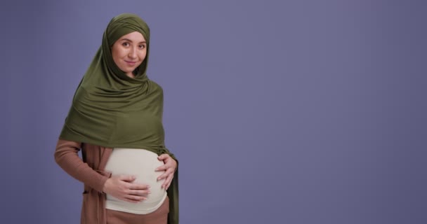 Szczęśliwa muzułmanka w ciąży pieści swój wielki brzuch, uśmiechając się do kamery na fioletowym tle z pustą przestrzenią — Wideo stockowe