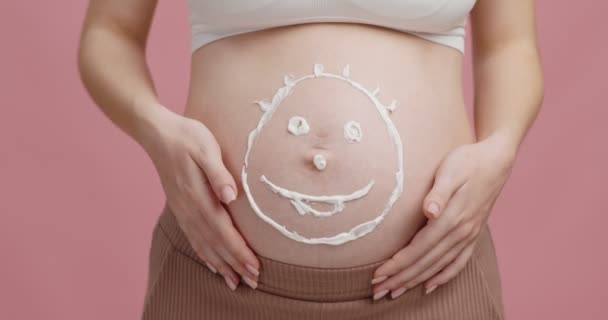 Graviditet och hudvård. Närbild av smiley ansikte ritad med grädde på stor mage, rosa studio bakgrund, slow motion — Stockvideo