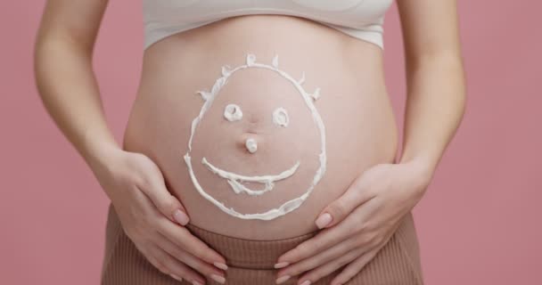 Cuidado de belleza para embarazadas. Señora expectante acariciando gran vientre con la cara sonriente crema dibujada, de cerca, fondo rosa — Vídeos de Stock