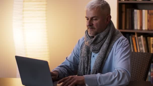 Zieke senior man gewikkeld in sjaal typen op laptop thuis, niezen en zich onwel voelen, slow motion — Stockvideo