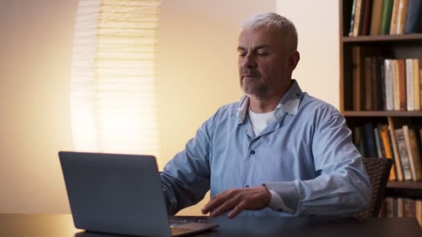 Hombre de mediana edad escribiendo en el ordenador portátil, estirando la espalda y seguir trabajando, necesita descansar, sentado en el lugar de trabajo en casa — Vídeos de Stock