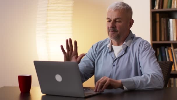 Седовласый мужчина средних лет машет рукой ноутбуку, прощается с коллегами и закрывает компьютер, сидит дома — стоковое видео