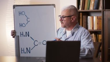 Çevrim içi kimya. Olgun profesör öğrencilere kimyasal formülleri açıklıyor, tahtaya yazıyor ve dizüstü bilgisayarına gösteriyor.