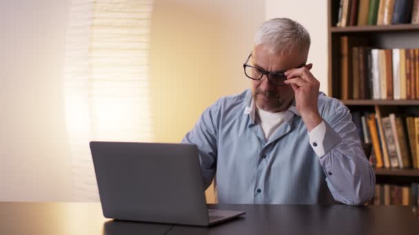 Проблемы со зрением. Зрелый мужчина снимает очки и массирует глаза, чувствуя усталость от работы на ноутбуке дома — стоковое видео