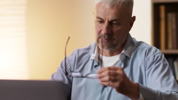 Ritratto ravvicinato dell'uomo anziano che si toglie gli occhiali e lo pulisce con uno straccio di microfibra, lavorando sul portatile a casa — Video Stock