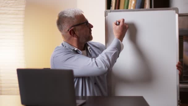 Profesor matematyki w średnim wieku pisze na tablicy, pokazując i wyjaśniając formuły do studentów online za pośrednictwem połączenia wideo — Wideo stockowe