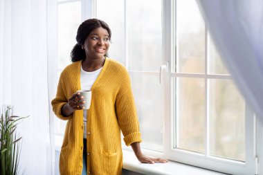 Sıradan giyinmiş, pencerenin yanında çay içen siyah bir kadın.