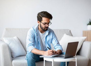 Çevrimiçi Eğitim. Doğu 'lu genç adam evde dizüstü bilgisayarla ders çalışıyor, not alıyor.