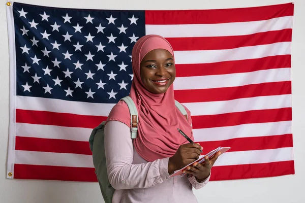 Smiling woman in hijab taking notes over american flag