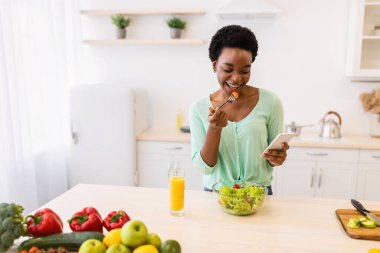 Siyah kadın cep telefonu kullanıyor, salata yiyor, mutfakta yemek yiyor.