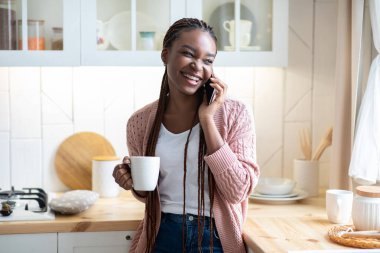 Neşeli siyah kadın cep telefonuyla konuşuyor ve mutfakta kahve içiyor.