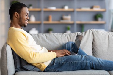 Kanepede dizüstü bilgisayar kullanarak uzanan siyah adamın yan görüntüsü